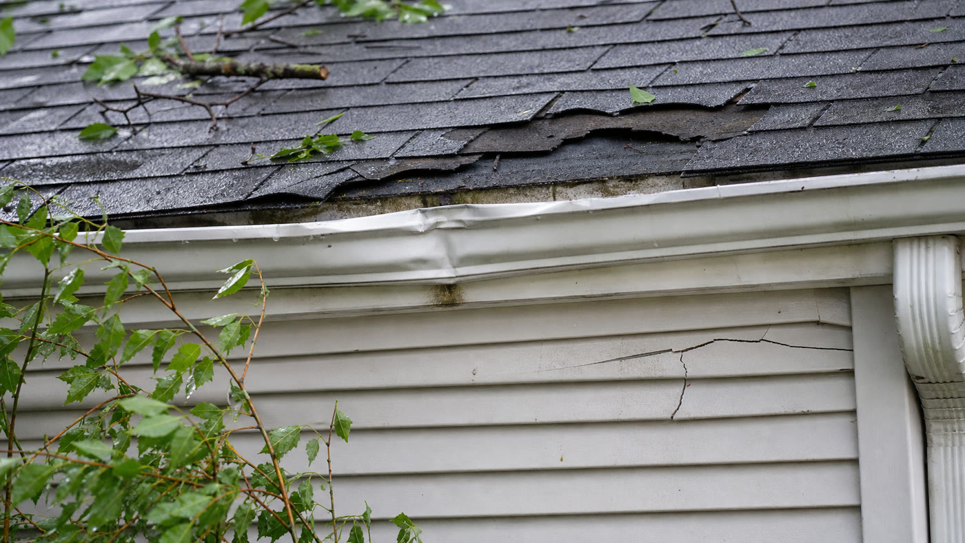 Lifted roof shingles, dented gutter, cracked siding, and storm debris on a home exterior